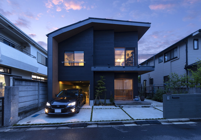 「三鷹のガレージハウス・東京」外観夕景を見る。木調の外壁材とオーダーメイドの木製玄関扉、ガレージ内は深いグリーンにする等、和のテイストでまとめられている。 設計／田中ノリヒト